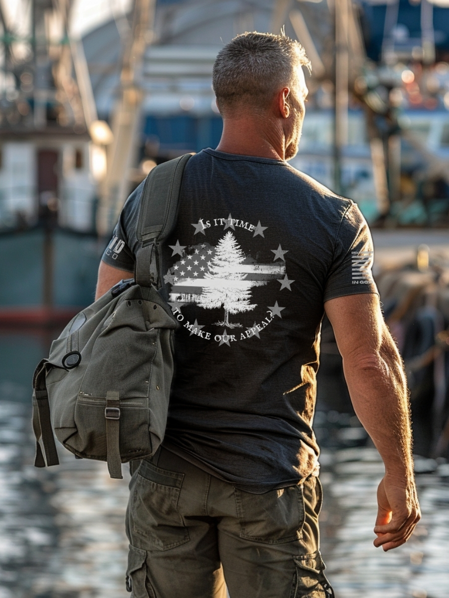 Model wearing dark heather grey t-shirt with Appeal to Heaven flag design featuring a bold White Pine Tree over a distressed American flag background. Historic patriotic graphic with strong revolutionary symbolism.