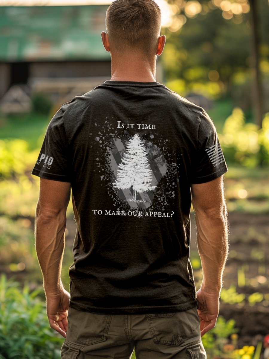 Model wearing black t-shirt with Appeal to Heaven flag design featuring a bold White Pine Tree over a distressed American flag background. Historic patriotic graphic with strong revolutionary symbolism.